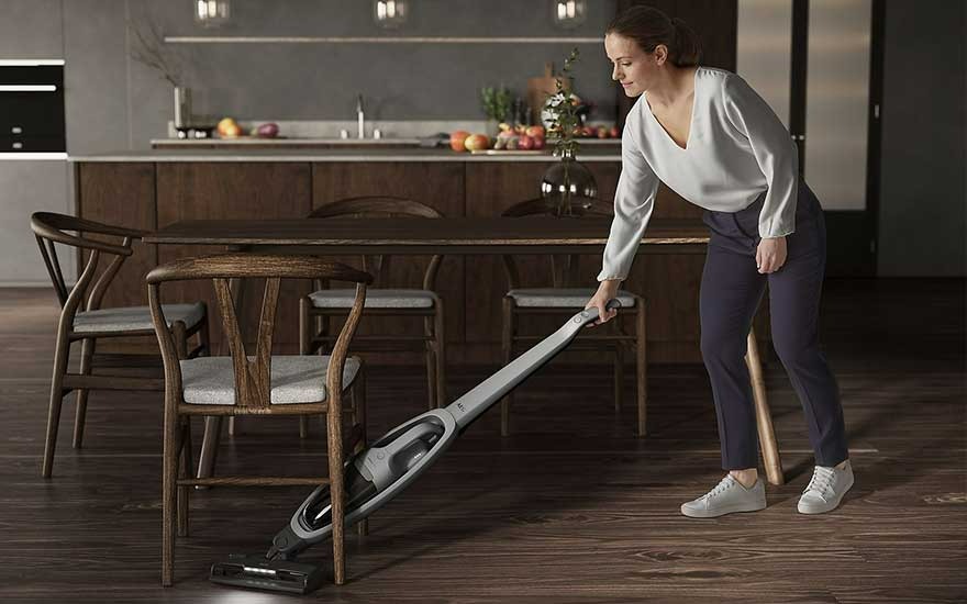 Une cuisine avec une table en bois et des chaises. Un aspirateur gris est utilisé sur le plancher en bois.