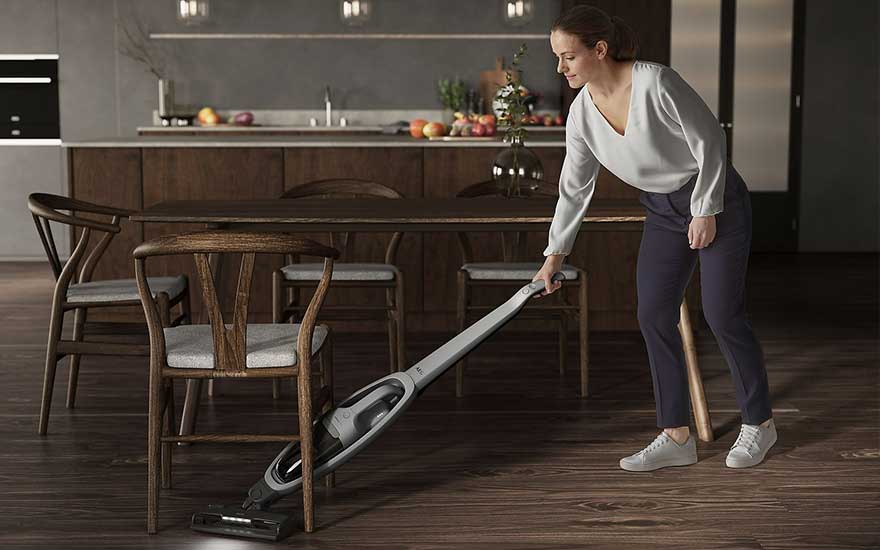 Een grijze stofzuiger staat op een houten vloer in een kamer met houten stoelen en een tafel.