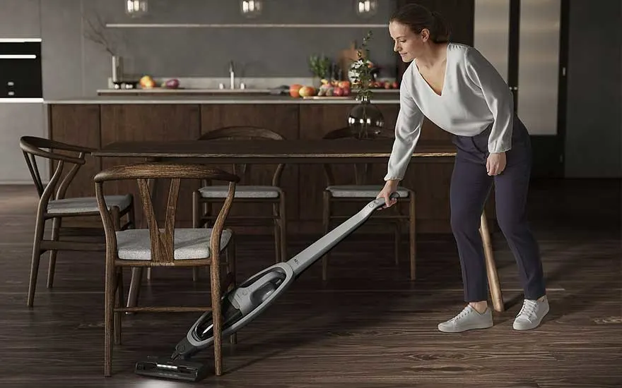 Een grijze stofzuiger staat op een houten vloer in een kamer met houten stoelen en een tafel.