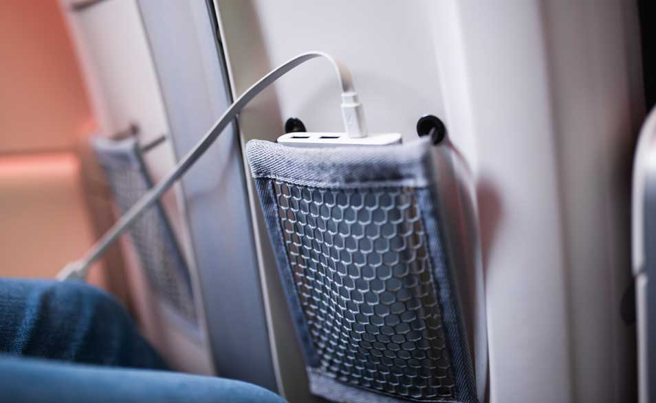 Powerbank in vliegtuigstoelzak, met witte kabel die naar buiten loopt, terwijl passagiersbeen ernaast rust in de krappe beenruimte.