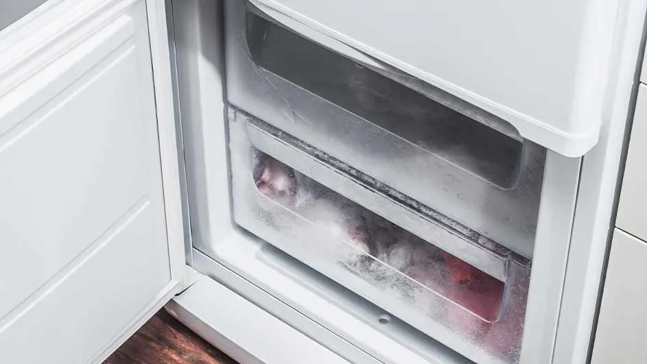 Un réfrigérateur blanc ouvert montre des tiroirs de congélation en plastique transparent, dont un avec des aliments congelés.