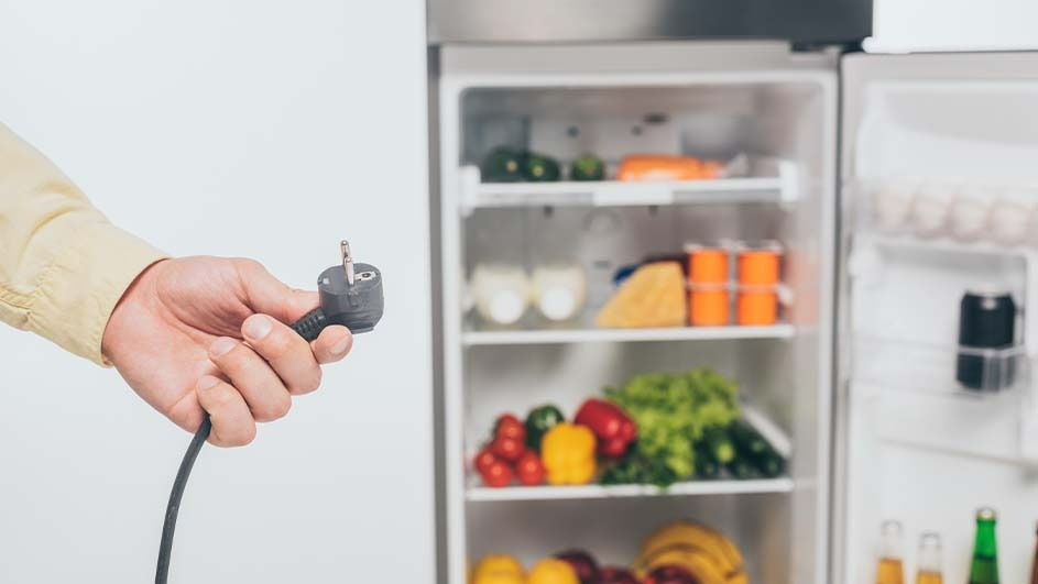 Une main tient une prise électrique noire devant un réfrigérateur blanc rempli de divers aliments colorés.