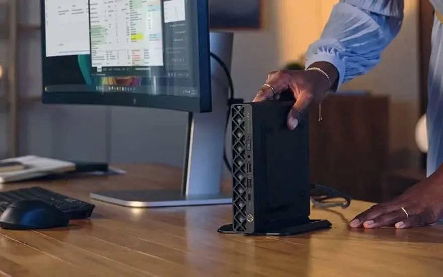 Een persoon plaatst een compacte HP-desktopcomputer naast een monitor op een houten bureau met toetsenbord en muis.