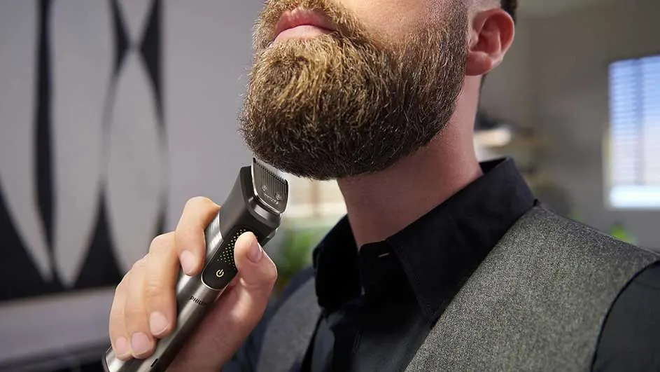 Een zilverkleurige trimmer wordt gebruikt om een bruine baard te scheren, met een grijze achtergrond.