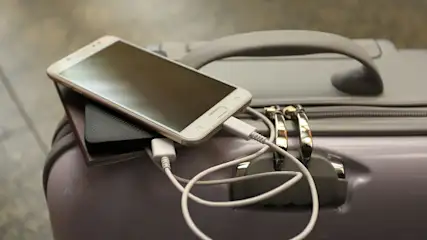 Een witte smartphone ligt op een zwarte powerbank op een grijze koffer met een wit oplaadsnoer.