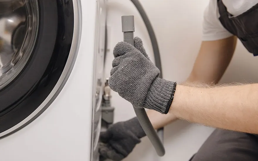 Een grijze slang wordt aangesloten op een witte wasmachine met een zwarte rubberen rand, met grijze gehandschoende handen.