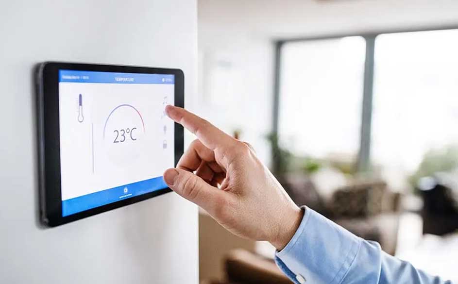 Een hand bedient een zwart tablet aan een witte muur, met een scherm dat 23°C toont, in een lichte kamer.