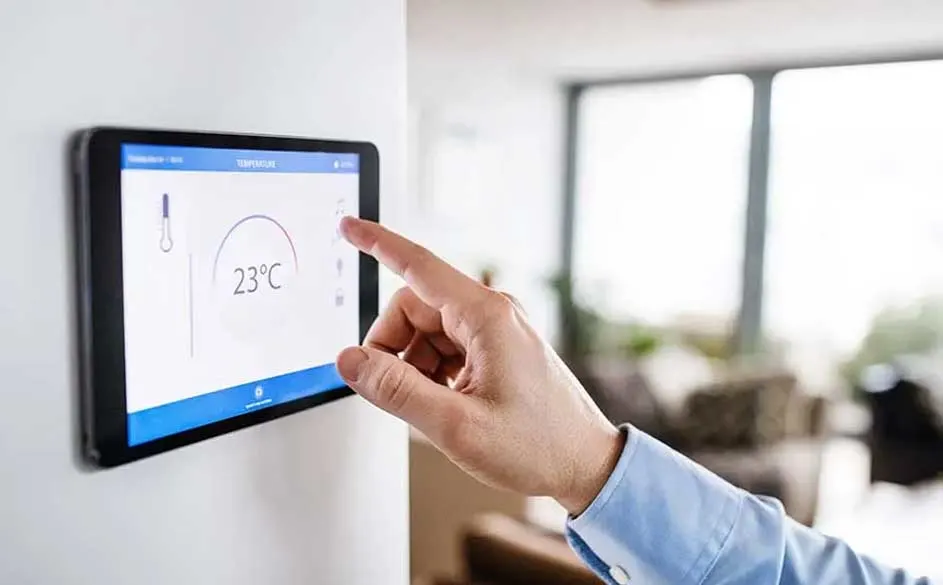 Een hand bedient een zwart tablet aan een witte muur, met een scherm dat 23°C toont, in een lichte kamer.