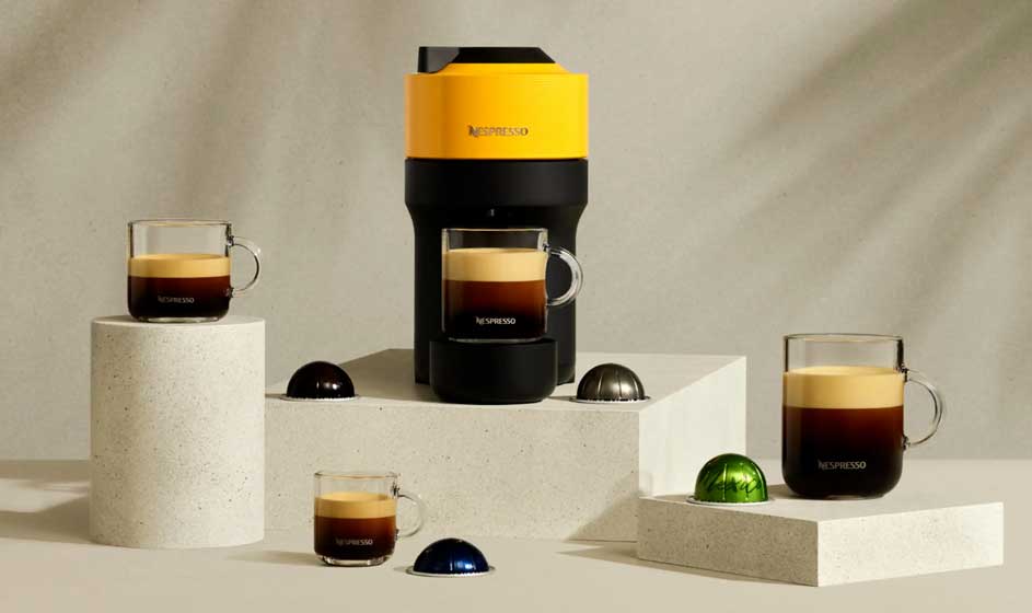 Een geel-zwarte Nespresso-machine staat op witte blokken, omringd door glazen koffie en kleurrijke capsules op een beige achtergrond.