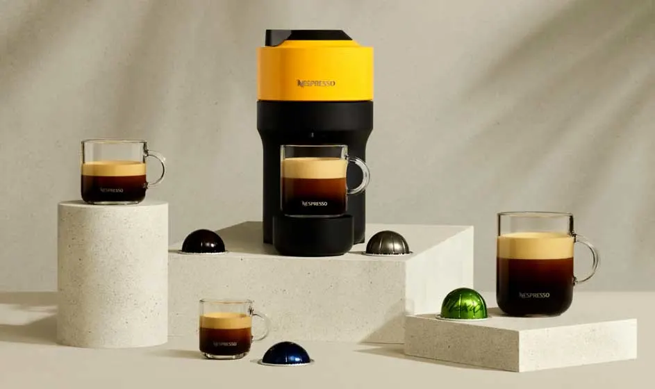 Een geel-zwarte Nespresso-machine staat op witte blokken, omringd door glazen koffie en kleurrijke capsules op een beige achtergrond.