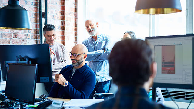 Kantoorinterieur met computers, een bakstenen muur en lampen. Er staan verschillende mensen in de kamer.