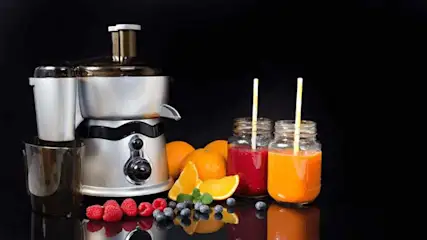 Zilveren juicer naast sinaasappels, frambozen, blauwe bessen en twee glazen met rode en oranje drank met rietjes.