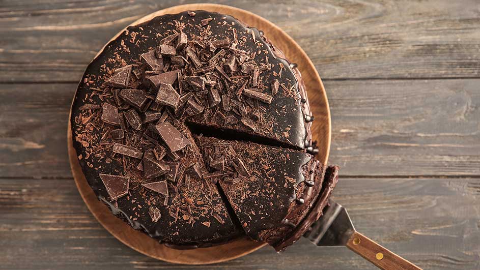 Een chocoladetaart met stukjes chocolade op een houten bord, een stuk wordt opgeschept met een taartschep.