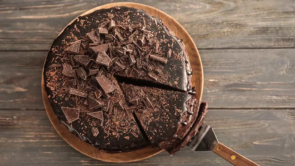 Een chocoladetaart met stukjes chocolade op een houten bord, een stuk wordt opgeschept met een taartschep.