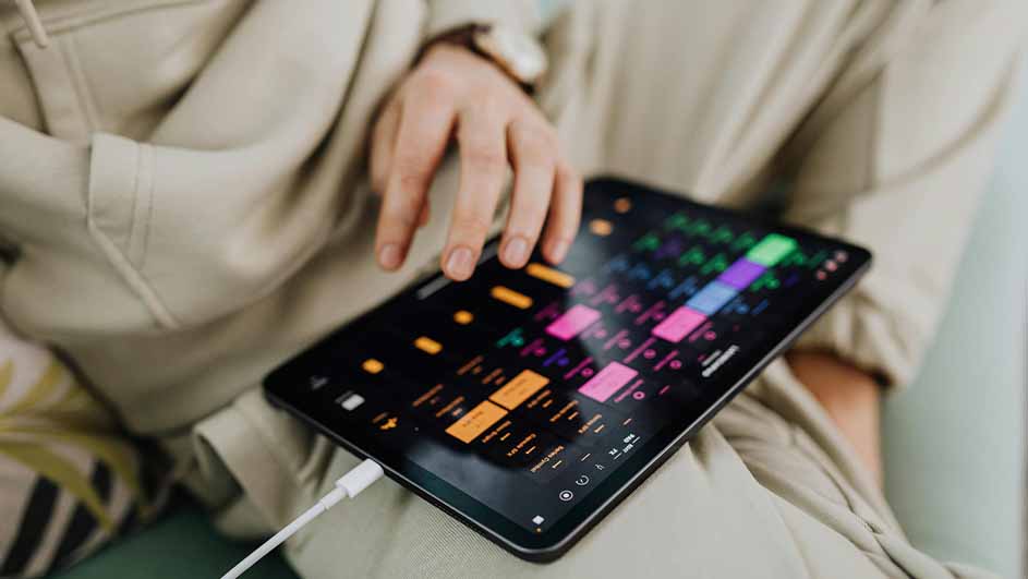 Un iPad noir affiche une application de musique colorée, avec un câble blanc branché en bas.