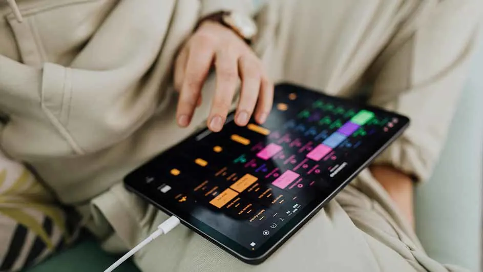 Un iPad noir affiche une application de musique colorée, avec un câble blanc branché en bas.