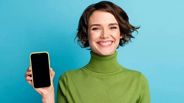 Une main tient un smartphone à écran noir avec une coque jaune, devant un fond bleu uni.