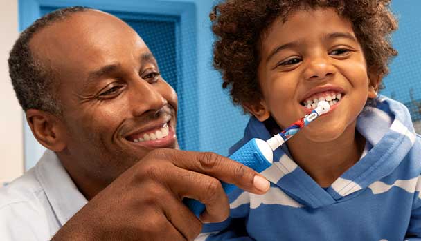 Un père aide son fils à se brosser les dents avec une brosse pour enfant; ils sourient, fond bleu et pull rayé visibles.