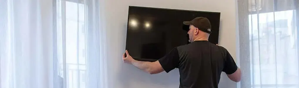 A flat-screen TV is being mounted on a wall, with white curtains and windows in the background.