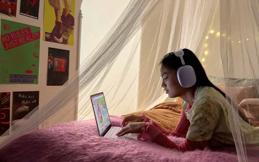 Young person lies on a pink bed with large white headphones, typing on a small laptop, surrounded by posters and warm fairy lights.