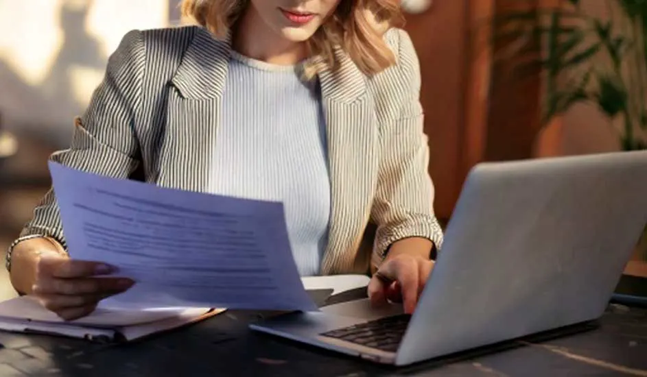 Een laptop en documenten op een tafel. Iemand draagt een gestreepte blazer en gebruikt de laptop.