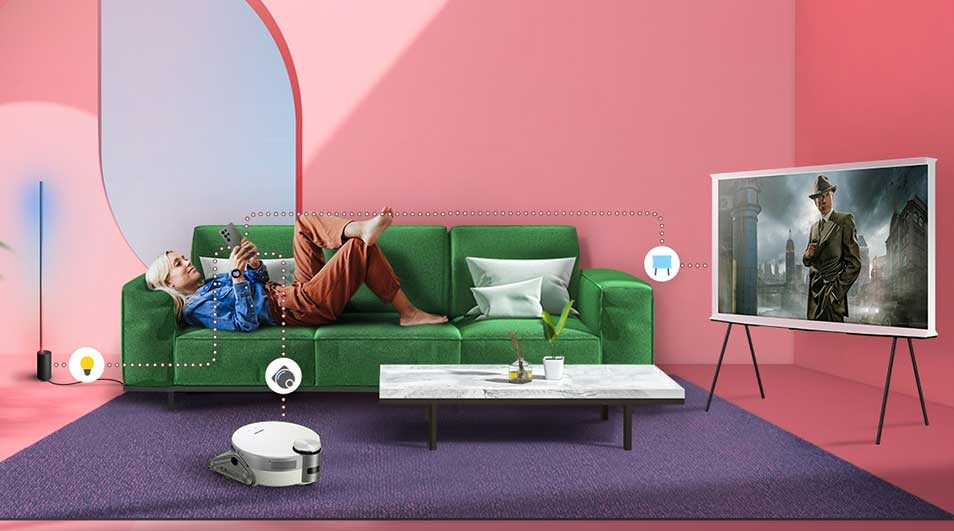 Salon rose avec canapé vert, tapis violet, table basse, téléviseur sur trépied et robot aspirateur blanc.