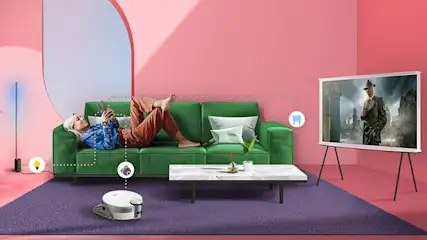 Salon rose avec canapé vert, tapis violet, table basse, téléviseur sur trépied et robot aspirateur blanc.