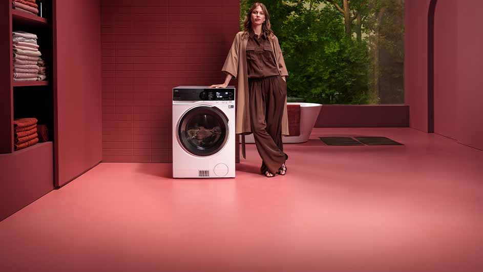 Een witte wasmachine in een roze kamer met een bad en groene planten op de achtergrond.