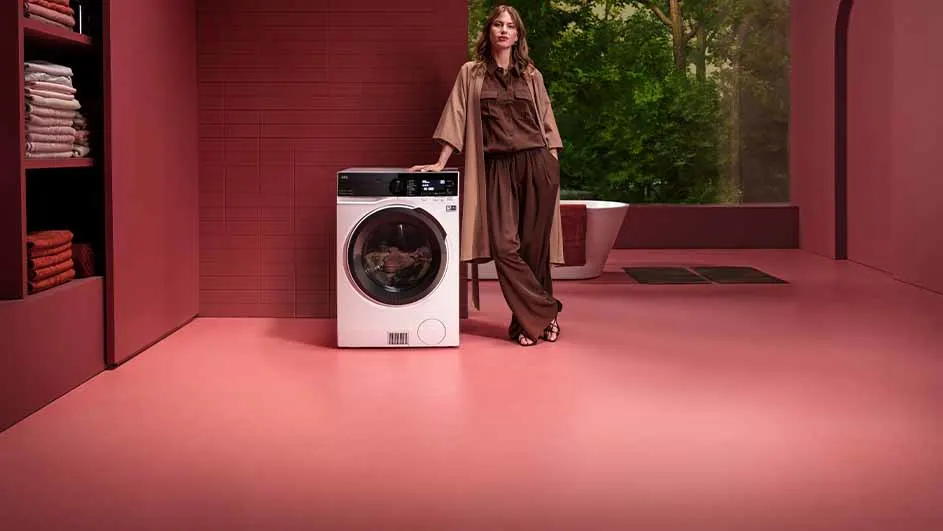 Un lave-linge blanc dans une pièce rose. Une étagère avec des serviettes est visible. À l'arrière, un bain et des arbres.