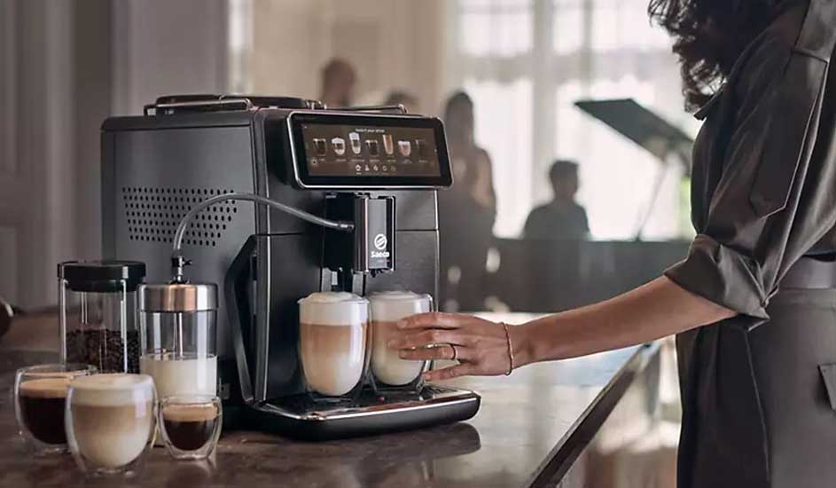 Une machine à café Saeco prépare des cappuccinos. Une femme place un verre, entourée de lait et de café dans un espace élégant.