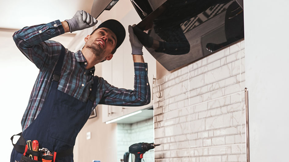 A person in work clothes is installing a black kitchen hood on a brick wall. Tools and a drill are visible.