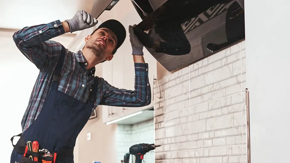 A person in work clothes is installing a black kitchen hood on a brick wall. Tools and a drill are visible.