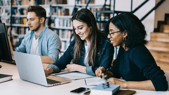 Trois personnes sont assises à une table avec un ordinateur portable, un ordinateur et des livres dans une bibliothèque.