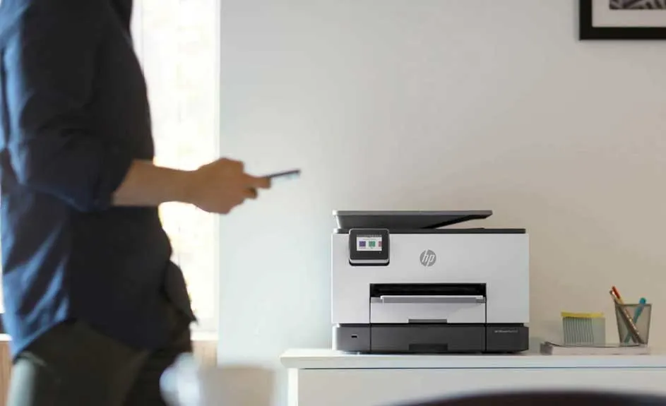Een grijze HP-printer staat op een wit bureau, een gsm wordt bediend, op een witte achtergrond.