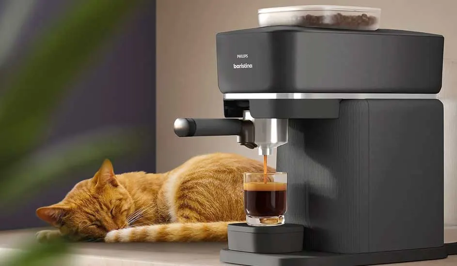 An orange cat sleeps beside a black espresso machine on a worktop. The machine is making coffee, with a small cup placed beneath the spout.