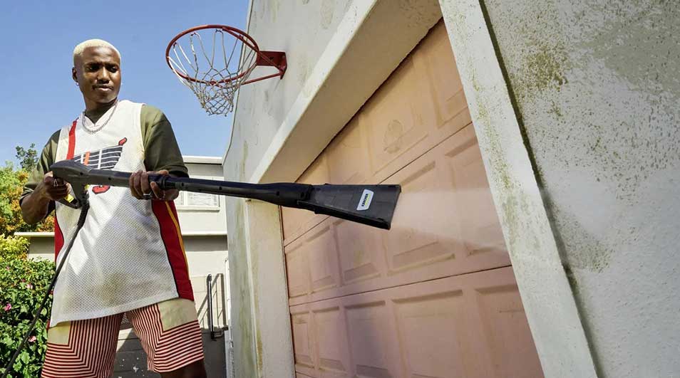 A person is cleaning the concrete patio slabs with a high-pressure washer whilst wearing rubber boots, in front of a contemporary house with a garden.