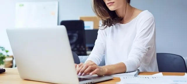 Une table en bois clair avec un ordinateur portable ouvert, des papiers et un stylo. Un écran blanc et une chaise noire sont visibles en arrière-plan.