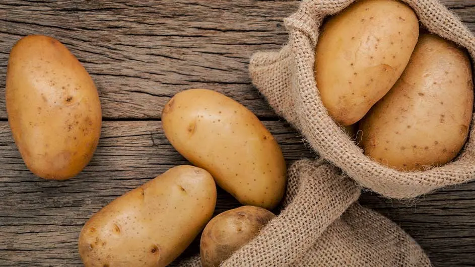 Op een houten tafel liggen aardappelen, waarvan een deel in een jute zak zit.