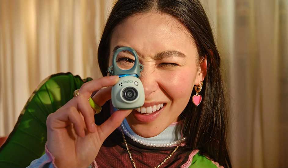 Une personne porte un haut vert, des bijoux dorés et tient une photo, avec de longs cheveux foncés et des rideaux clairs en arrière-plan.