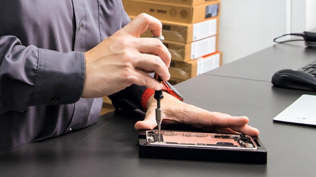 Un tournevis répare un téléphone noir sur un bureau gris. En arrière-plan, des boîtes sont empilées.