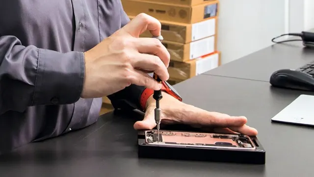 Un tournevis répare un téléphone noir sur un bureau gris. En arrière-plan, des boîtes sont empilées.