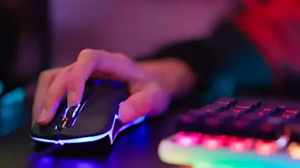 Hand bedient een zwarte gamingmuis met blauwe LED-verlichting. Een toetsenbord met rode en groene verlichting is zichtbaar op de achtergrond.