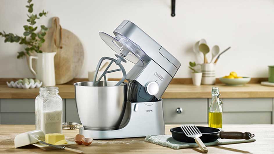 Un robot de cuisine argenté avec bol en acier, sur un comptoir de cuisine avec ingrédients et ustensiles.