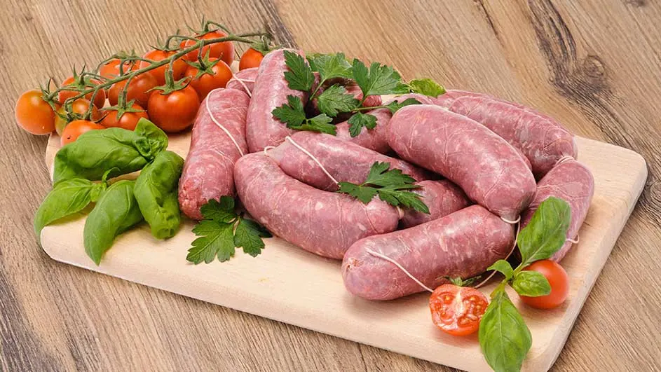 Saucisses crues sur une planche à découper en bois, avec tomates cerises rouges et feuilles vertes de basilic et de persil.