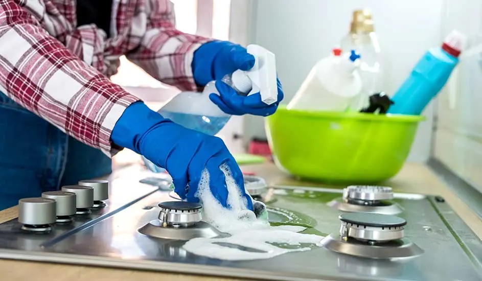 Une personne nettoie une cuisinière en acier inoxydable avec des gants bleus, du savon et un vaporisateur. Un bac vert contient des produits.