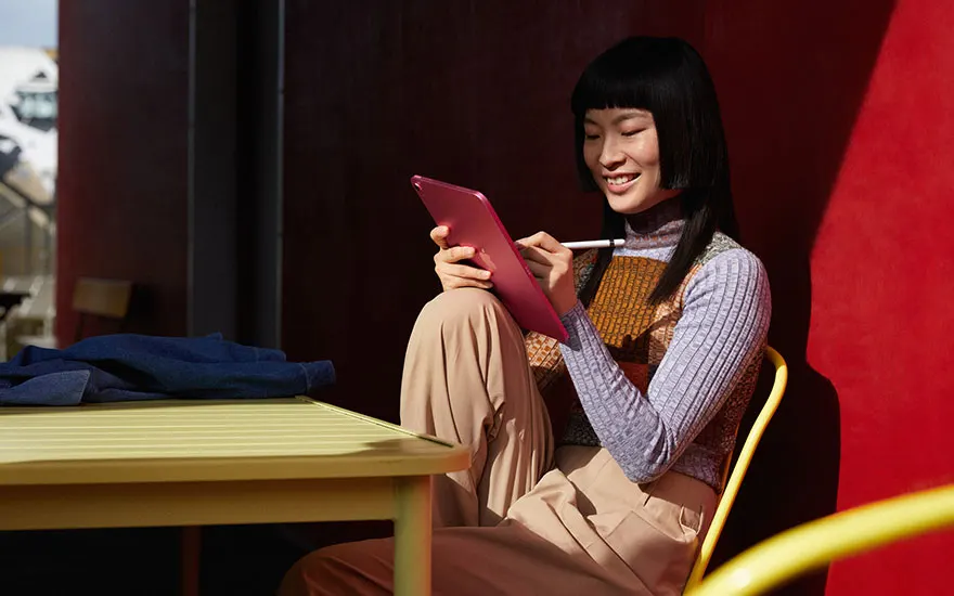 Woman with black hair sits relaxed on yellow chair using red iPad in modern space with colourful red background setting.