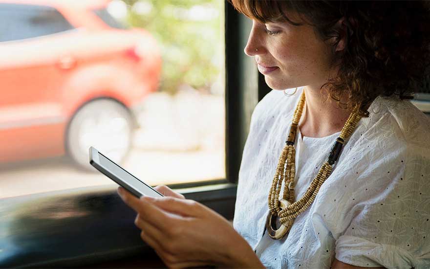 Une personne tient une tablette devant une fenêtre. Une voiture orange est visible à l'extérieur. Elle porte un collier à plusieurs rangs.