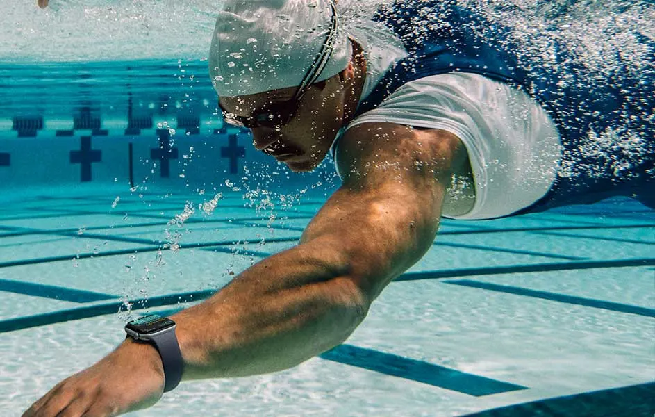 Een zwemmer duikt krachtig door het heldere zwembadwater. Hij draagt een badmuts, zwembril en smartwatch, terwijl luchtbellen opstijgen.