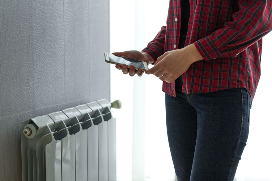Un mur gris jouxte un radiateur blanc. Une personne en chemise à carreaux rouge tient un smartphone.
