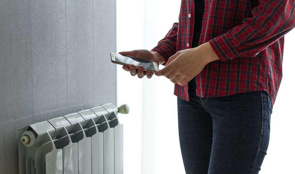 Een grijze radiator staat naast een grijze muur. Iemand houdt een telefoon vast, draagt een rood geruit hemd en donkere jeans.
